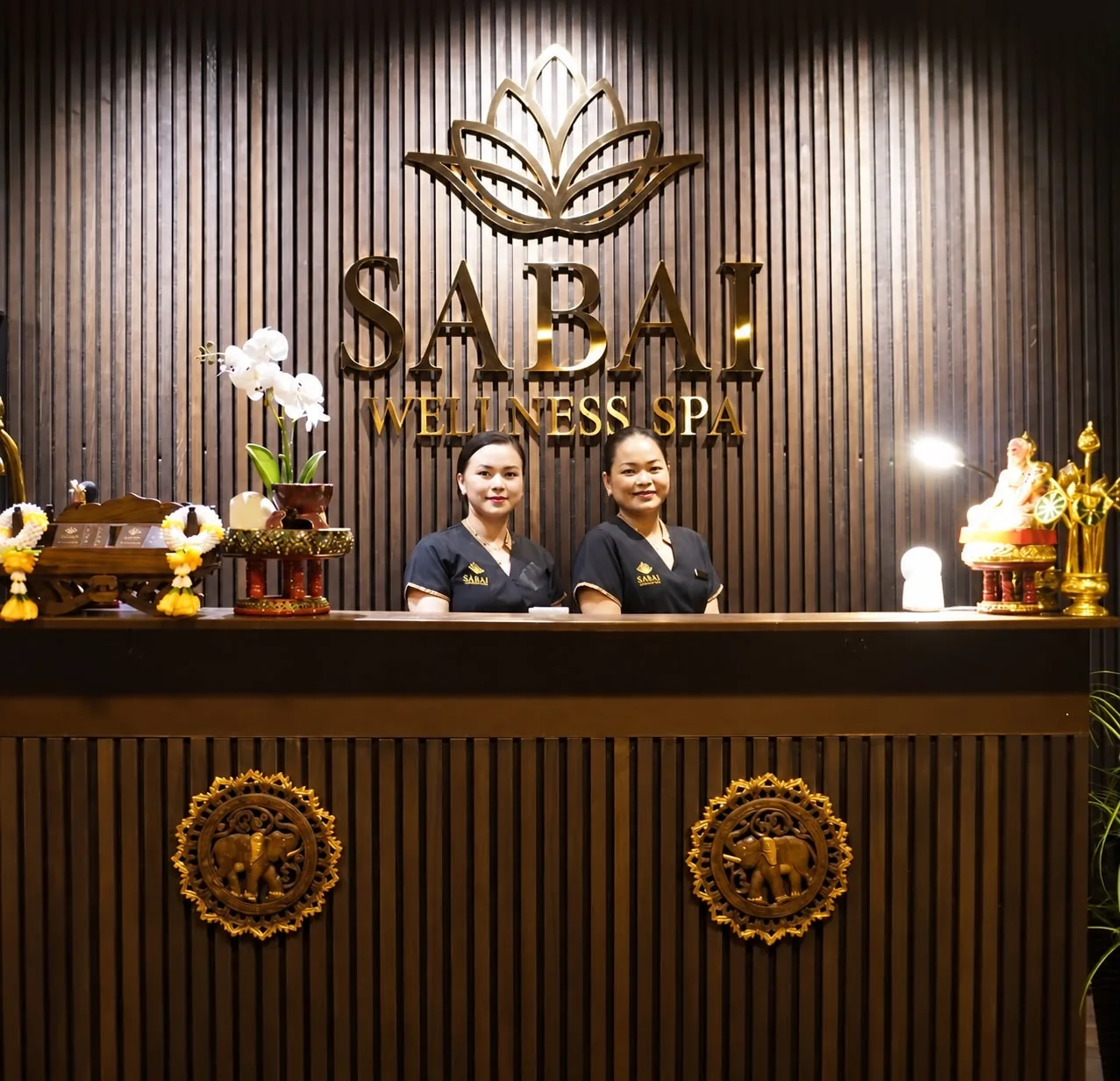 Kaksi Sabain työntekijää vastaanottotiskin takana, seinällä kultainen SABAI WELLNESS SPA -logo